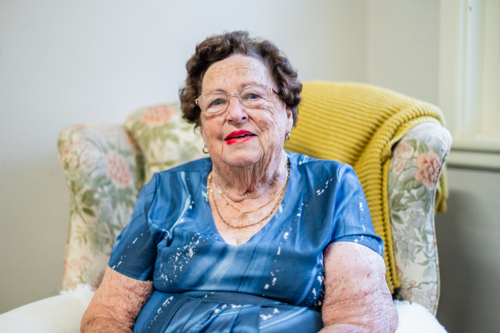 Portrait of elderly woman in a blue dress sitting on a patterned armchair - Australian Stock Image