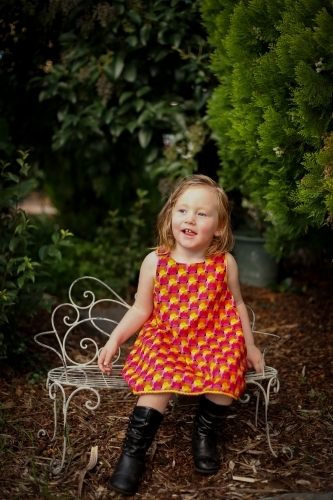Portrait of female child wearing bright red dress sitting in natural garden setting - Australian Stock Image