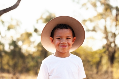 Portrait of happy aboriginal boy wearing hat for sun protection with bokeh bushland background - Australian Stock Image