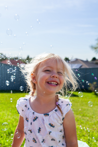Portrait of little girl with joyful smile outdoor surrounded by bubbles in Australian backyard - Australian Stock Image