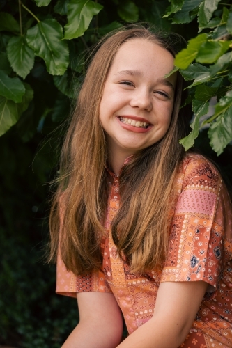 Portrait of pre-teen girl sitting on brick wall with natural green leafy background - Australian Stock Image