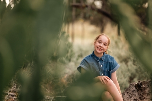 Portrait of pre-teen girl sitting on log in natural setting with big smile - Australian Stock Image