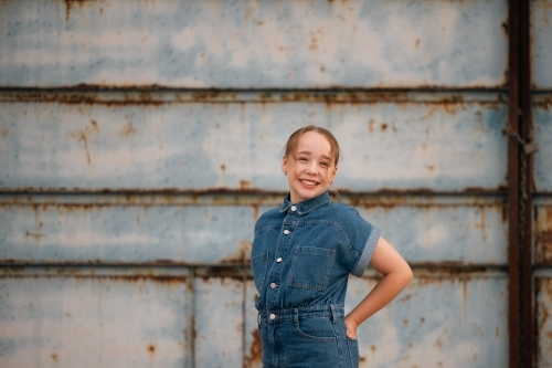 Portrait of pre-teen girl wearing denim jump suit in front of large blue barn door - Australian Stock Image