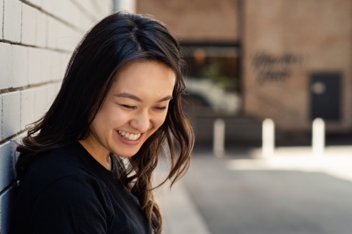 portrait of smiling asian woman, casual portrait leaning against a brick wall - Australian Stock Image