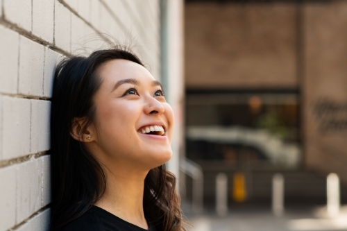 portrait of smiling asian woman, casual portrait leaning against a brick wall - Australian Stock Image