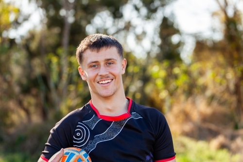 Portrait of smiling teen First Nations footy player with ball in Australia - Australian Stock Image