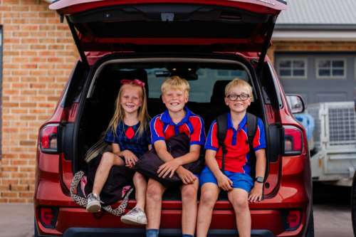 portrait of three siblings sitting together in car boot ready for school drop off smiling - Australian Stock Image