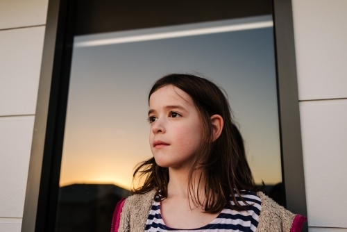 Portrait of young girl watching the sunset which is reflected in a house window behind her