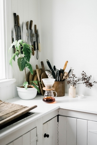 Pour over coffee on kitchen bench with utensils, knives and plants behind.