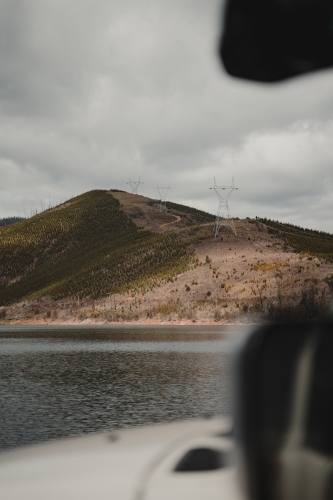 Powerlines on a mountain through a pine forest clearing with a lake in the foreground - Australian Stock Image