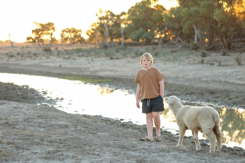 Pre-teen boy walking along creek being followed by pet sheep in dry remote bush setting - Australian Stock Image