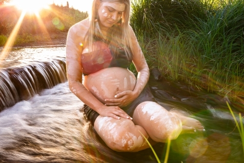 Pregnant aboriginal woman sitting in flowing water bonding with her unborn child at sunset - Australian Stock Image