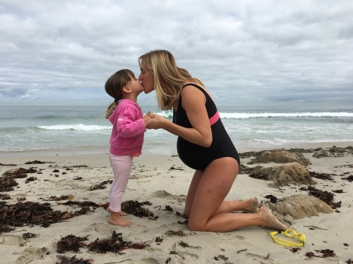 Pregnant mother kissing toddler daughter at beach on overcast day