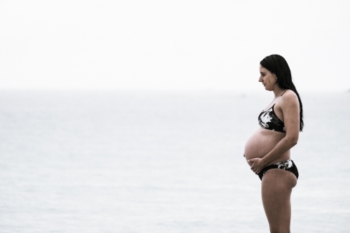 pregnant woman on beach in bikini holding onto belly on overcast rainy day mid shot