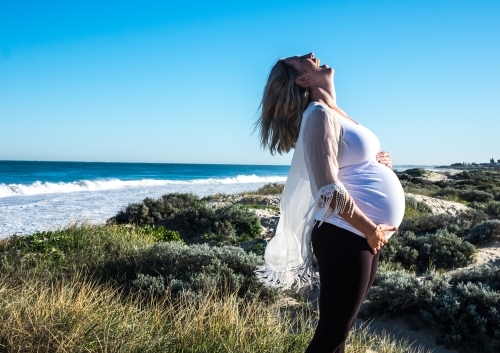 Pregnant woman standing in sand dunes with ocean background - Australian Stock Image