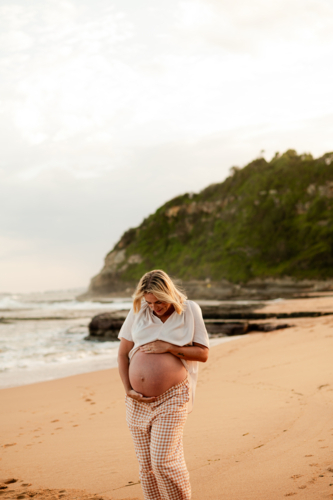 Pregnant woman walking along a sandy beach at sunset with gentle waves nearby - Australian Stock Image