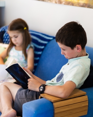Primary age siblings reading books together on the lounge - Australian Stock Image