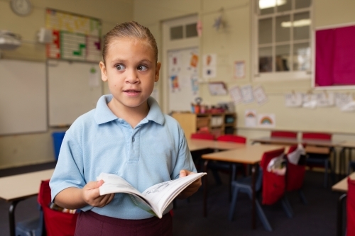 Primary school girl student standing holding a book in a classroom - Australian Stock Image
