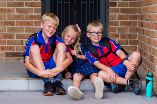 Primary school students sitting together on front doorstep of home ready to go to school - Australian Stock Image