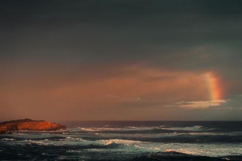 Rainbow above cliffs with waves at glowing sunset - Australian Stock Image