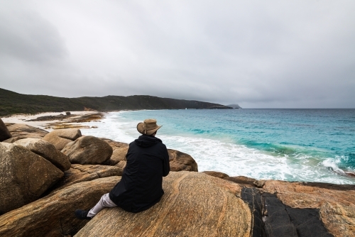Rear view of a person sitting on a rock looking out at sweeping view of coastline with stormy sky - Australian Stock Image