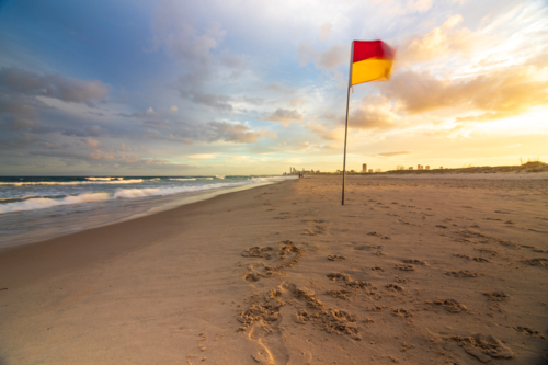 Red and yellow lifeguard flag blowing in wind on sandy beach with tide out and sunset sky - Australian Stock Image