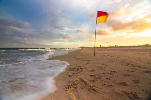 Red and yellow lifeguard flag on sandy beach with waves crashing - Australian Stock Image