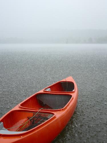 Red canoe sits idle on grey rainswept lake.