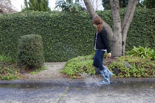 Red haired girl splashing in puddles in gumboots on a rainy winter day.