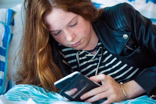 Red haired teenager girl lying on her bed while playing on her phone. - Australian Stock Image