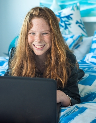 Red haired teenager working on the computer while lying on the bed. - Australian Stock Image