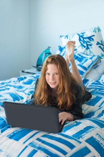 Red haired teenager working on the computer while lying on the bed. - Australian Stock Image