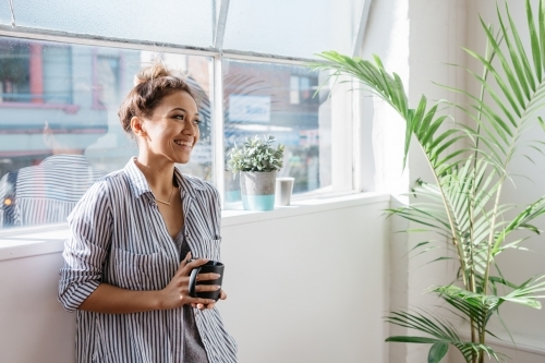 Relaxed female employee standing with a cup of coffee by the window - Australian Stock Image
