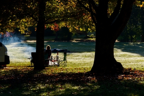 Rim lit silhouette of person sitting at picnic table with smoke under trees with autumn colours - Australian Stock Image