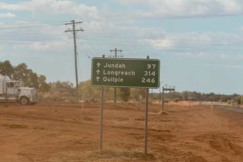Road sign on dusty, red roadside with parked truck in background.