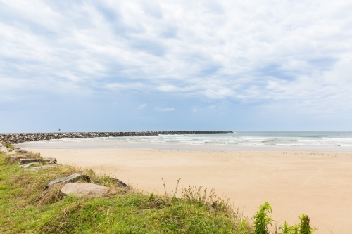 Rock wall curving around beach with sand and water and cloudy skies - Australian Stock Image