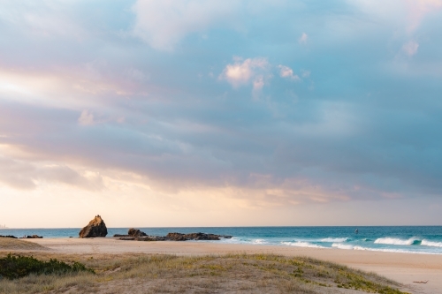 Rocks on Currumbin Beach illuminated at sunset with person windsurf foiling in the distance - Australian Stock Image