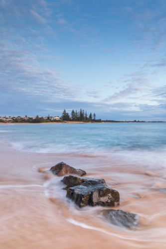 Rocks on golden sand surrounded by ocean with headland in background - Australian Stock Image