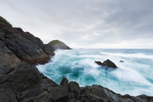 Rough ocean waves crashing against black rocks on an overcast day - Australian Stock Image