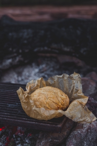 Rustic damper bread in brown baking paper on the red hot coals of a campfire - Australian Stock Image