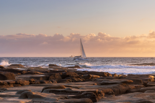 Sailboat on ocean with rocky foreshore and cloudy sky - Australian Stock Image