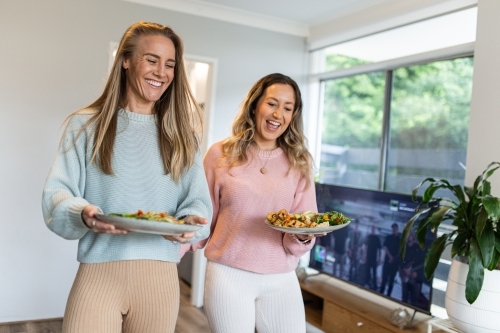 Same sex couple carrying their plates of food together at home - Australian Stock Image