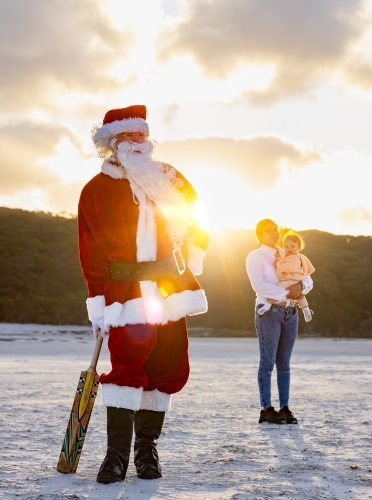 santa claus on the beach with a cricket bat with young woman and baby looking on - Australian Stock Image