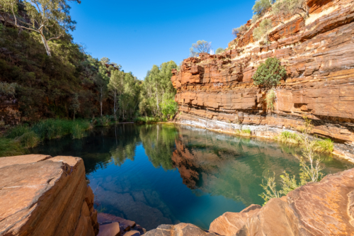Scenic natural rock pool surrounded by red cliffs and lush vegetation under a clear blue sky. - Australian Stock Image
