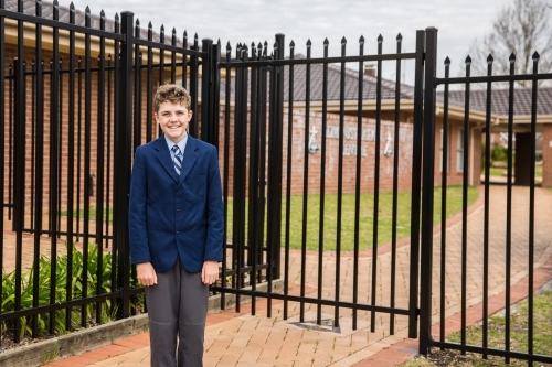School aged boy in uniform standing at entry to school near security fence - Australian Stock Image
