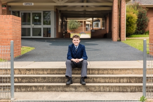 School child in uniform sitting on stairs in front of school office smiling - Australian Stock Image