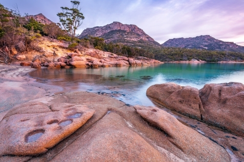 Sculptured granite rocks around a still bay with large mountains behind at twilight - Australian Stock Image