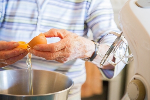 Senior citizen cooking a cake in the kitchen, cracking eggs into a bowl - Australian Stock Image
