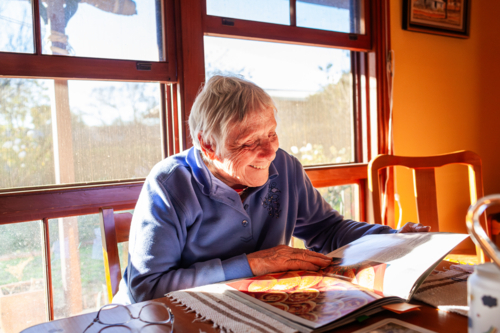 Senior woman reading cook book deciding what to bake sitting in home in winter sunshine - Australian Stock Image
