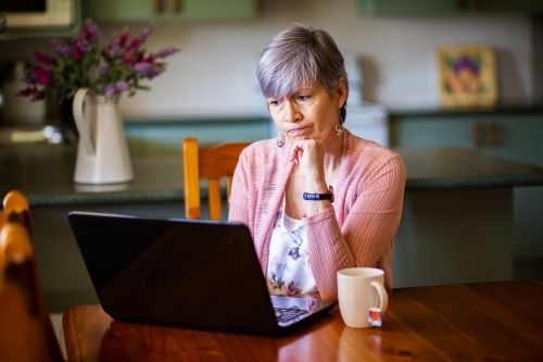 Senior woman with cup of tea working on laptop from home - Australian Stock Image
